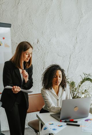 Two businesswomen sharing ideas on a project, working together on a laptop in a modern office setting.