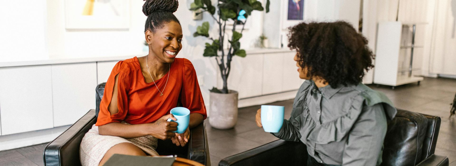 Two businesswomen enjoy a coffee break and conversation in a modern office setting.