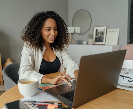 A woman sits at a round table, working on a laptop with a coffee cup nearby in a cozy home office setting.